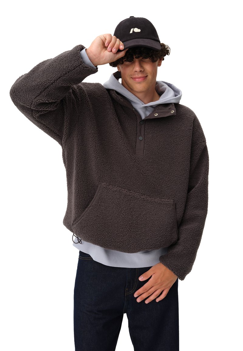 Brązowy polarowy pulower z kieszenią z przodu, z zapięciem na napy, nałożony na szary rozpinany podkoszulek, noszony z ciemnymi dżinsami i czarną czapką.