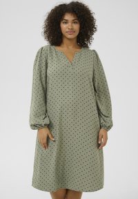 Robe verte avec une coupe décontractée, décolleté en V et longues manches bouffantes. Présente un motif à pois noirs sur un tissu texturé.