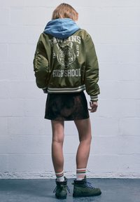 Giacca bomber verde con grafica di una tigre, felpa con cappuccio blu sotto, gonna in suede nera e stivali da trekking verdi con lacci blu.