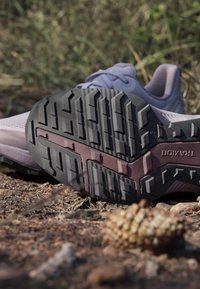 Nahaufnahme der Schuhsohle mit grobem Profil auf einem unbefestigten Weg, die das Gripmuster und die Marke "TRAXION" zeigt.