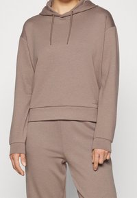 Sudadera con capucha y corte corto en suave tejido beige con puños acanalados y cordón ajustable. Presenta un sutil logo de la marca cerca del dobladillo.