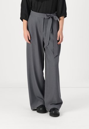 Person trägt graue, hoch taillierte, weit geschnittene Hose mit gebundenem Gürtel, schwarze Schuhe und ein schwarzes Langarmoberteil vor einem einfachen Hintergrund.