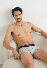 Boxers en coton gris avec une ceinture marine arborant le logo Lacoste, ornés de petits logos bleus, portés par un modèle assis sur une chaise.