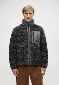 Fleece bunda s textúrovaným čiernym a šedým vzorom, vysoký golier, plný zips na prednej strane a zipsová náprsní vrecko v šedej syntetickej látke.