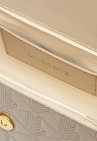 Intérieur d'un sac à main beige avec un motif géométrique, présentant une texture lisse, une doublure en satin et une étiquette avec le logo Lacoste sur un fond clair.