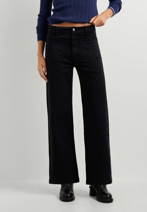 Donna che indossa pantaloni neri a vita alta e gamba larga, stivaletti neri e maglione a costine blu navy con maniche lunghe.