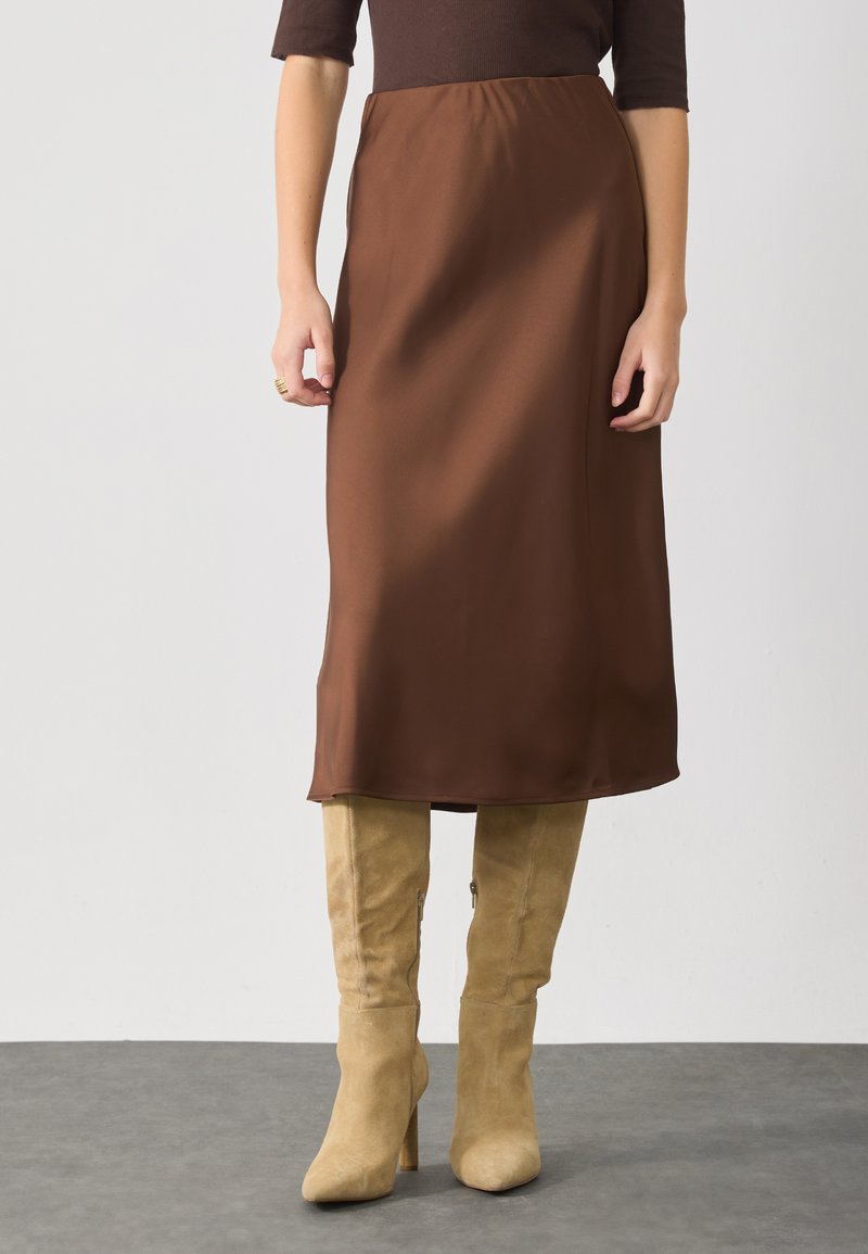 Brauner Satin-Midirock mit elastischem Bund, kombiniert mit beigen, hochhackigen Wildlederstiefeln. Glatte Textur, schlichtes Design.