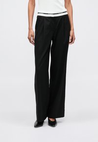 Čierne nohavice s širokými nohavicami a skladmi, so snehobielym brandovaným pásom. Hladká textúra a strih vhodný na mieru, doladené s čiernymi špicatými topánkami.