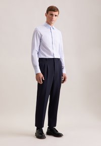 Camicia a maniche lunghe azzurro chiaro con una sottile trama, abbinata a pantaloni pieghettati blu scuro e scarpe nere con lacci. Posizione in piedi.