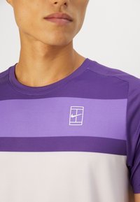 Nærbilde av en person iført en lilla, lyslilla og hvit stripete Nike sportsdrakt med logo på brystet.