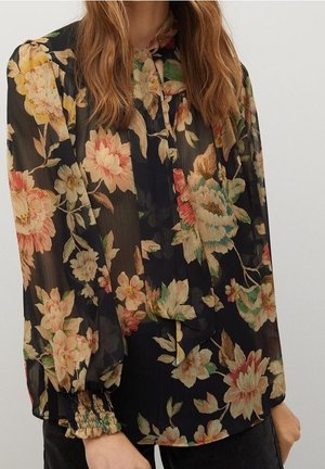 Czarna przezroczysta bluzka z dużym różowym, beżowym i zielonym kwiatowym wzorem, długie, szerokie rękawy i marszczone mankiety, noszona z ciemnymi spodniami.