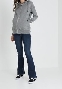 Persona incinta che indossa un cappuccio grigio con zip, jeans svasati blu scuro e sneakers nere, in piedi su uno sfondo bianco uniforme.