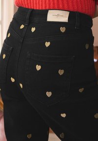 Jeans negri cu inimioare mici brodate în aur și etichetă "Love & Roses", purtate cu un pulover roșu din tricot.