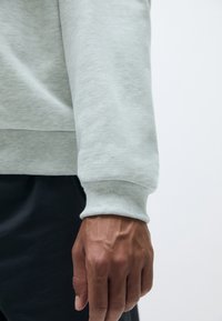 Manga de sudadera gris claro con puño acanalado, mostrando una textura suave. Una mano sostiene la manga, revelando venas detalladas.