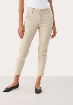 Woman wearing slim-fit beige ankle-length pants with side pockets and black pointed-toe flats against a plain background.