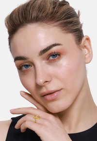 Anello d'oro con un design intrecciato, indossato su un dito. Il modello ha la pelle liscia, occhi blu e un'espressione neutra. Maglietta nera.