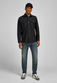 Zwarte lange mouwen knoopshirt met twee borstzakken, gecombineerd met lichtblauwe jeans en zwart-witte sneakers, en een zwarte pet.
