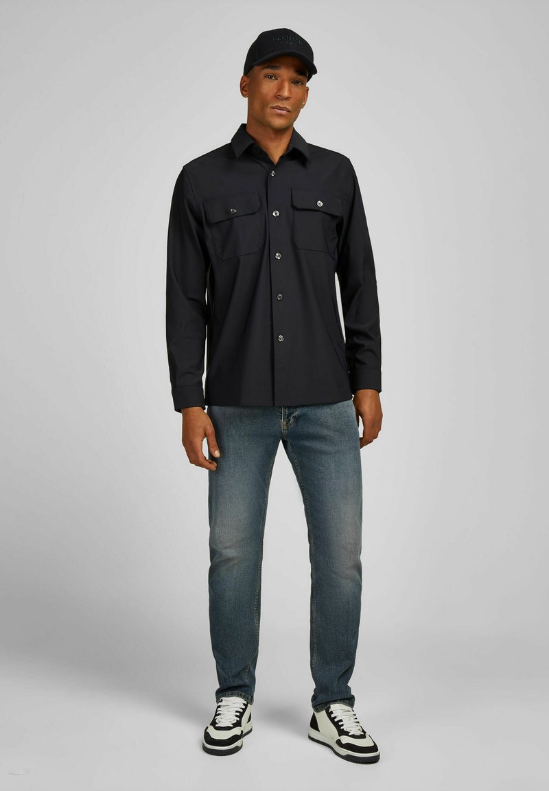 Zwarte lange mouwen knoopshirt met twee borstzakken, gecombineerd met lichtblauwe jeans en zwart-witte sneakers, en een zwarte pet.