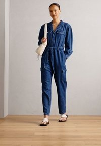 Jeansoverall med lång ärm, framfickor och midje bälte, i en medelblå färg, matchad med vita sandaler och en liten väska.