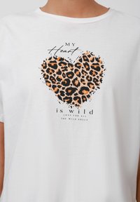 Balta kokvilnas t krekls ar leoparda apdrukas sirdi oranžā un melnā krāsā. Uz tā ir uzraksts: "MANAS sirds ir savvaļas mīlestība visiem SAVVAĻAS GARU cilvēkiem."