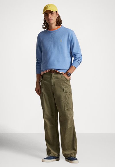 Φωτεινό μπλε πουλόβερ, πορτοκαλί ριγέ πουκάμισο από κάτω, χακί παντελόνι cargo, κίτρινο καπέλο και σκούρα μπλε παπούτσια. Υλικό από βαμβάκι, καθημερινό σχέδιο.