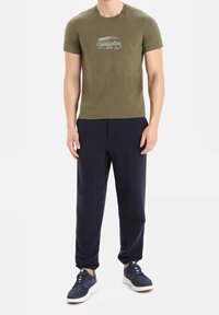 Olivgrön t-shirt med kort ärm och en grafik av en husvagn; kombinerad med marinblå sweatpants och marinblå sneakers med vita sålar.