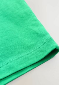 Nærbillede af grøn bomulds T-shirt-stof med en ensartet tekstur og kantede kanter, liggende fladt på en neutral baggrund.