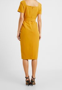 Senapsgul klänning med fyrkantig halsringning, korta ärmar och markerad midja. Slät tyg, knälång, med dragkedja bak.