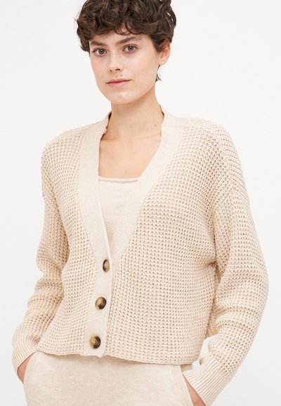 Beige cropped cardigan with a waffle knit texture, v-neck, and wooden buttons. Long sleeves with ribbed cuffs and relaxed fit.