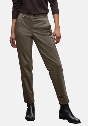 Person trägt olivgrüne gerade geschnittene Hose, dunkelbraues Langarmshirt und schwarze Stiefeletten und steht vor weißem Hintergrund.