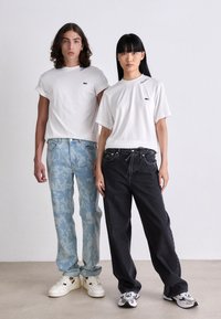 Dva modelové stojí vedle sebe, oba mají na sobě bílé trička Lacoste, na levé straně mají světlé vzorované džíny, na pravé volné černé džíny a bílé tenisky.