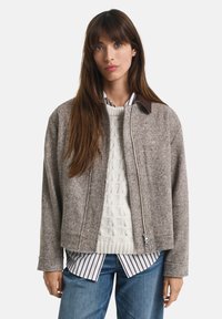 Woman wearing a gray zip-front jacket over a cream cable-knit sweater, striped shirt, and blue jeans, standing against a white background.