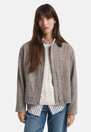 Woman wearing a gray zip-front jacket over a cream cable-knit sweater, striped shirt, and blue jeans, standing against a white background.