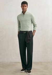 Šviesiai žalias megztinis su užtrauktuku, tamsios kelnės su juosmens raišteliu ir rudi sportbačiai. Švelnios tekstūros, laisvas siluetas, sluoksniuotas stilius.