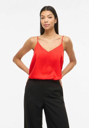Kvinde iført rød ærmeløs V-hals top og sorte bukser med høj talje, stående med hænderne bag ryggen mod en hvid baggrund.