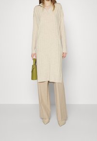 Ljust beige stickad klänning med krage, med vertikal ribbad textur; ihop med ljusbeige byxor och en liten grön handväska.