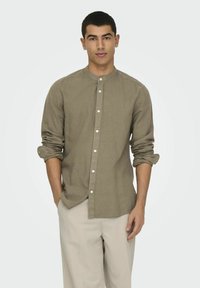 Camicia button-up color tan con colletto alla cinese, maniche lunghe arrotolate ai polsi, abbinata a pantaloni beige chiaro. Tessuto dal finish liscio.