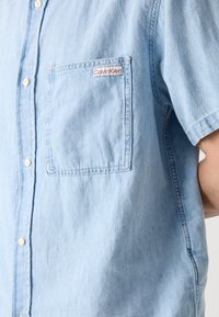 Camisa de manga corta azul clara hecha de tela de mezclilla, con un bolsillo con un logo en rojo de "Calvin Klein" y botones blancos a lo largo del frente.
