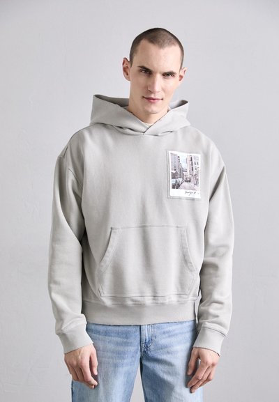 Haut à capuche gris clair avec une poche sur le devant, présentant un détail d'impression photographique. Coupe légèrement ample et texture douce, associé à un jean bleu clair.