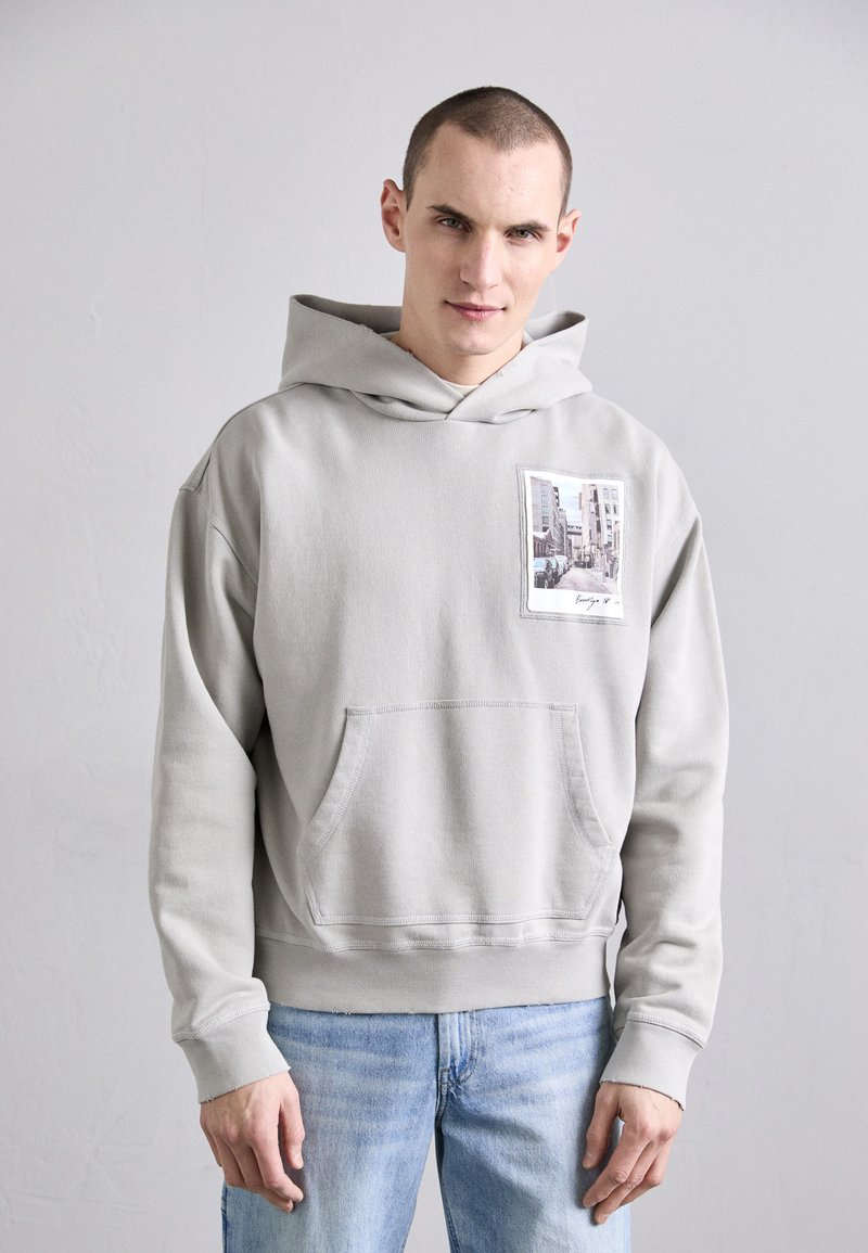 Jasnoszara bluza z kapturem z kieszenią z przodu, z detalem z nadrukiem fotograficznym. Lekko luźny krój i miękka tkanina, w połączeniu z jasnoniebieskimi dżinsami.