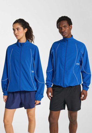 To modeller iført blå atletikjakker med hvid piping; den ene kombineret med marineblå shorts, den anden med sorte shorts, stående mod en hvid baggrund.