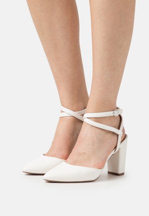 Chaussures à talons hauts blanches avec un bout pointu, une bride réglable à la cheville et un talon bloc épais. Matière en cuir lisse avec peu de détails.