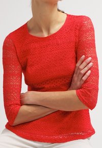 Haut en dentelle rouge avec des manches trois-quarts, présentant un motif en zigzag et un col rond. Le tissu est texturé et léger.