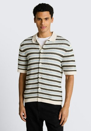 Junger Mann trägt ein beige-blau-braun gestreiftes, kurzärmeliges, geknöpftes Strickhemd über einem weißen T-Shirt, mit schwarzen Hosen.