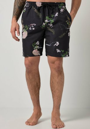 Schwarze Shorts mit floralem Muster, elastischem Bund und Kordelzug, getragen von einem stehenden Mann mit nackten Beinen und tätowiertem Arm.