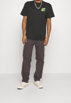 Homme portant un t-shirt graphique noir, un pantalon cargo foncé, des baskets blanches et des chaînes argentées sur un fond blanc uni.