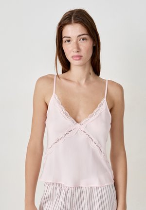Giovane donna con lunghi capelli castani che indossa un top in pizzo rosa chiaro e pantaloni del pigiama a righe, su uno sfondo semplice.