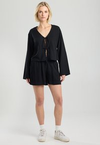 Musta lyhythihainen pusero, jossa on solmimisdetalji edessä ja pitkät hihat, yhdistetty mustiin shortseihin. Mallilla on valkoiset lenkkarit ja sukat. Silkkinen pinta.