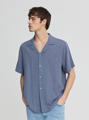 Giovane uomo con capelli castani corti che indossa una camicia blu a maniche corte e larghe con bottoni e jeans azzurri, che guarda verso sinistra.