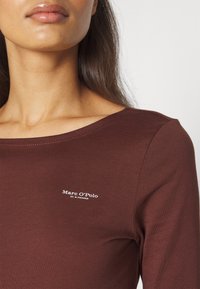 Brun langærmet top lavet af ribbet stof. Har en rund halsudskæring og et lille hvidt brandlogo på brystet.